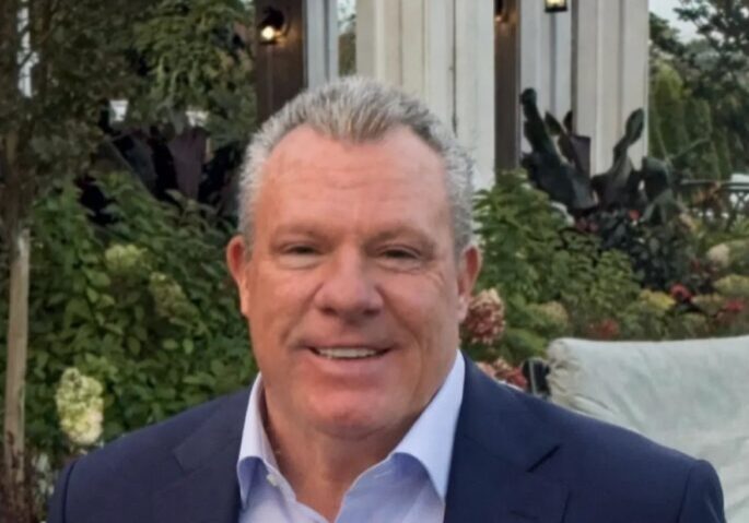 Smiling man in a navy blazer and light blue shirt outdoors.